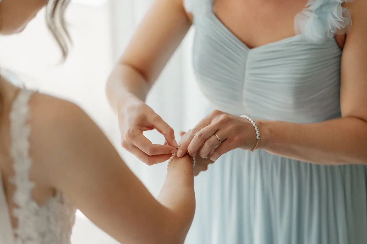Eine Frau im plissierten hellblauen Kleid mit Stoffblüten an der Schulter schließt ein Perlenarmband an Annas Handgelenk, an ihrem eigenen Arm ein Armband aus Buchstabenperlen.