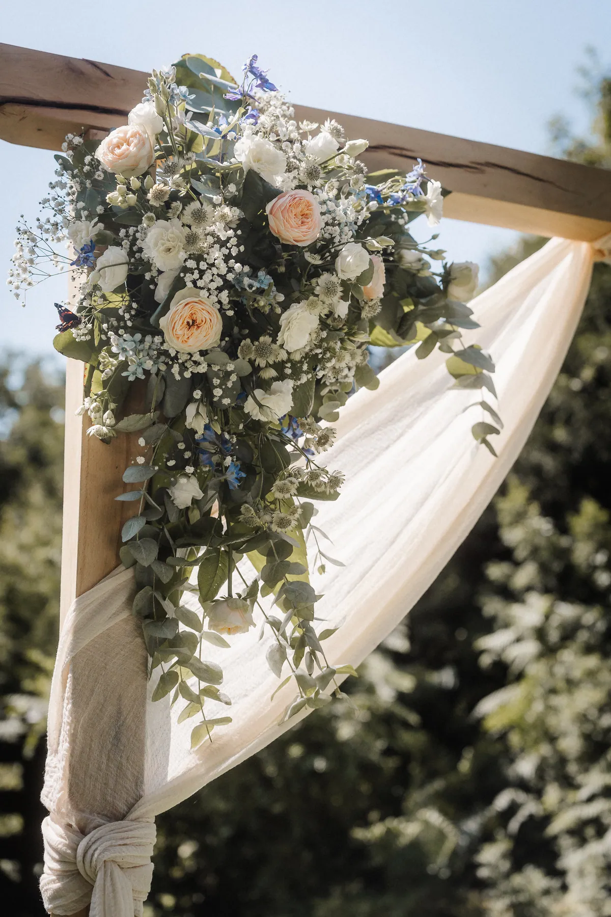 Eine Ecke des hölzernen Traubogens ist mit Rosen, Schleierkraut, blauen Blüten und Eukalyptus geschmückt, ein cremefarbenes Tuch ist um den Pfosten geknotet, links sitzt ein Schmetterling.