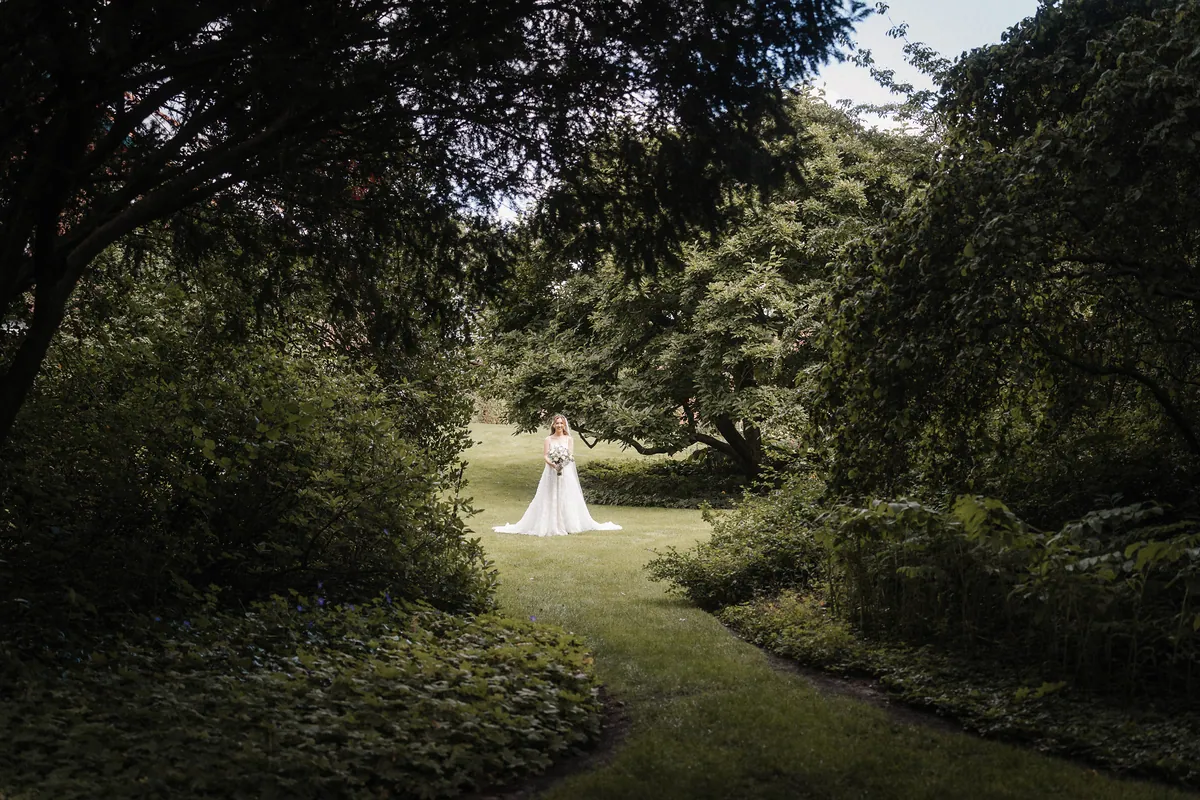 Dunkle Baumkronen und Büsche rahmen den Blick über einen geschwungenen Rasenweg zu Anna, die lächelnd im Brautkleid mit Schleier auf der hellen Wiese steht, den Strauß vor sich, die Schleppe im Gras aufgefächert.