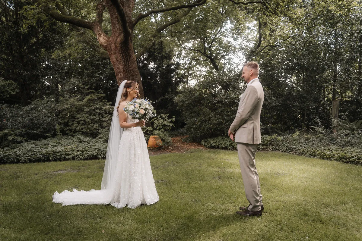 Anna und Kai stehen sich auf dem Rasen gegenüber und lächeln einander an, sie hält einen Strauß aus Pfirsichrosen, weißen und blauen Blüten, hinter ihr ein alter Baum.
