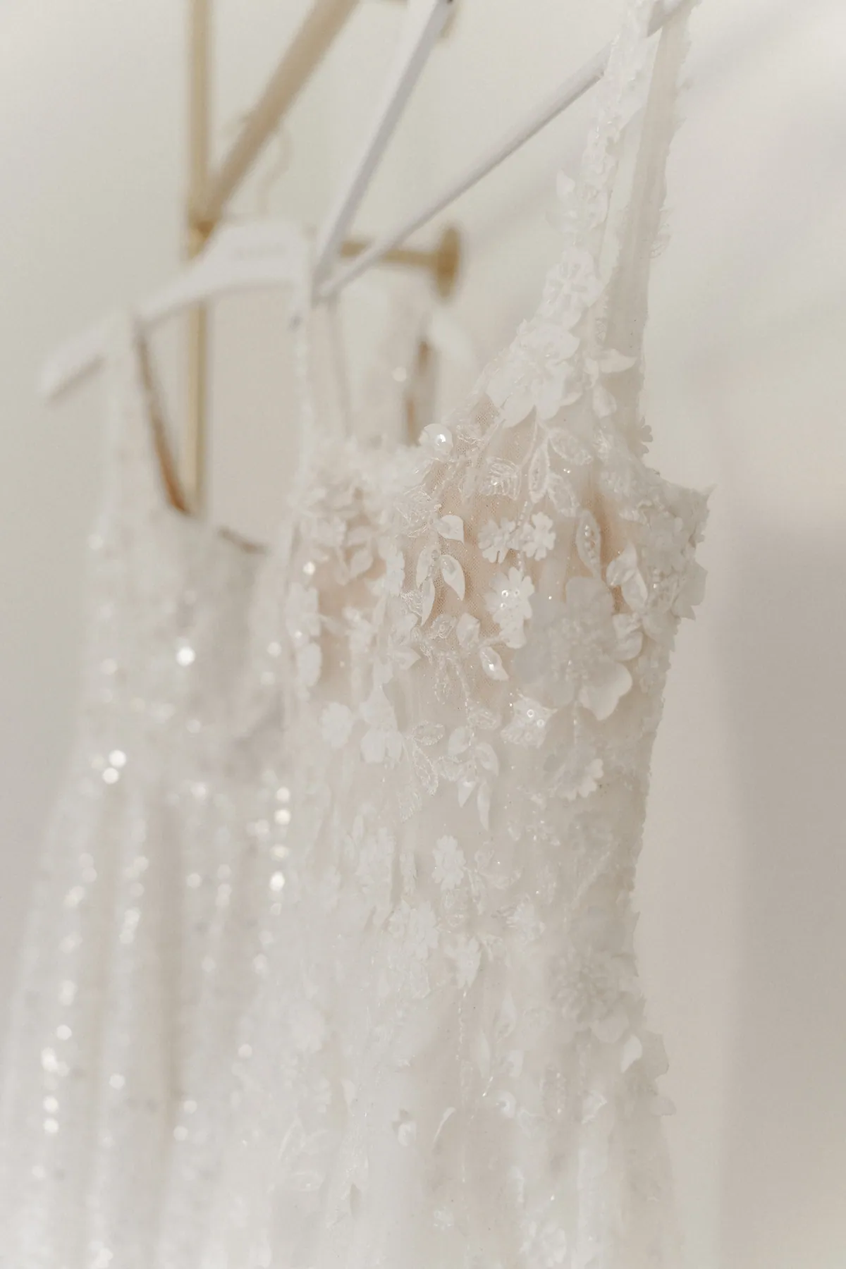 Nahaufnahme des Brautkleids am Bügel: plastische Spitzenblüten, Blätter und Pailletten auf zartem Tüll, dahinter unscharf ein zweites, glitzerndes Kleid an der goldenen Kleiderstange.