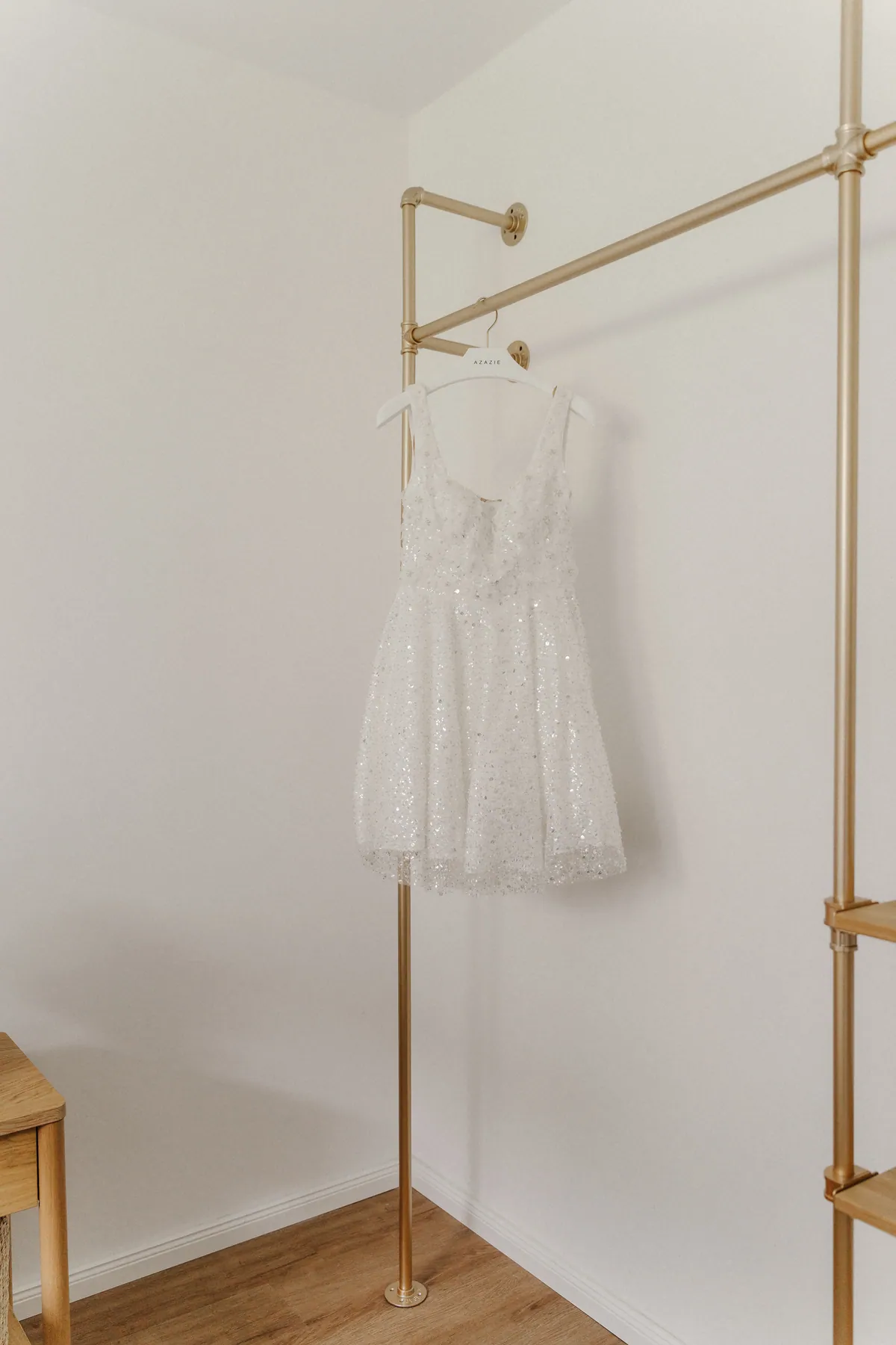 In einer hellen Zimmerecke hängt ein kurzes, über und über mit Pailletten besetztes weißes Kleid auf einem weißen Bügel an einer goldenen Rohrgarderobe über Holzboden.
