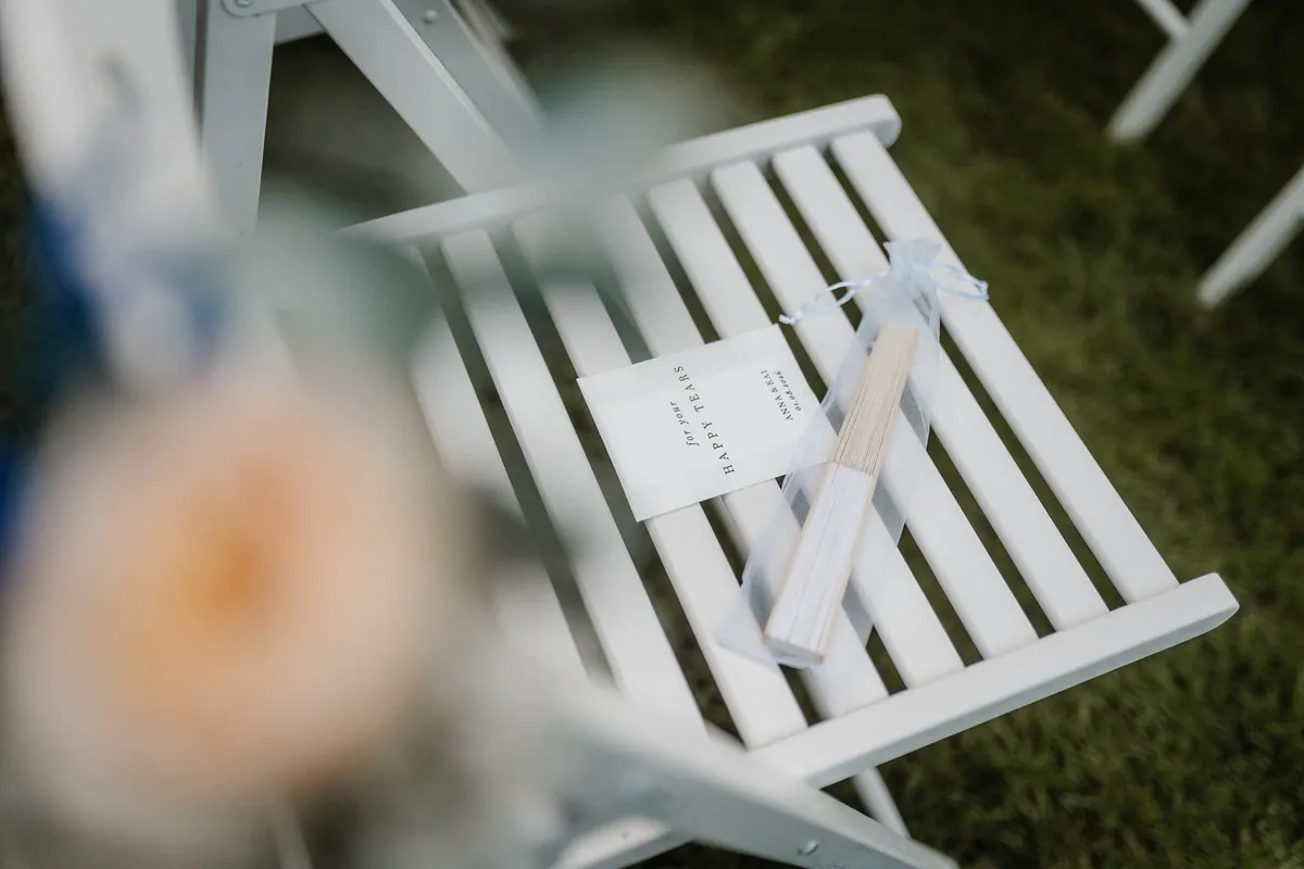 Auf einem weißen Klappstuhl liegen ein Kärtchen mit der Aufschrift „for your HAPPY TEARS – ANNA & KAI – 01.08.2026“ und ein Fächer im Organzabeutel, vorn unscharf Blüten eines Gestecks.