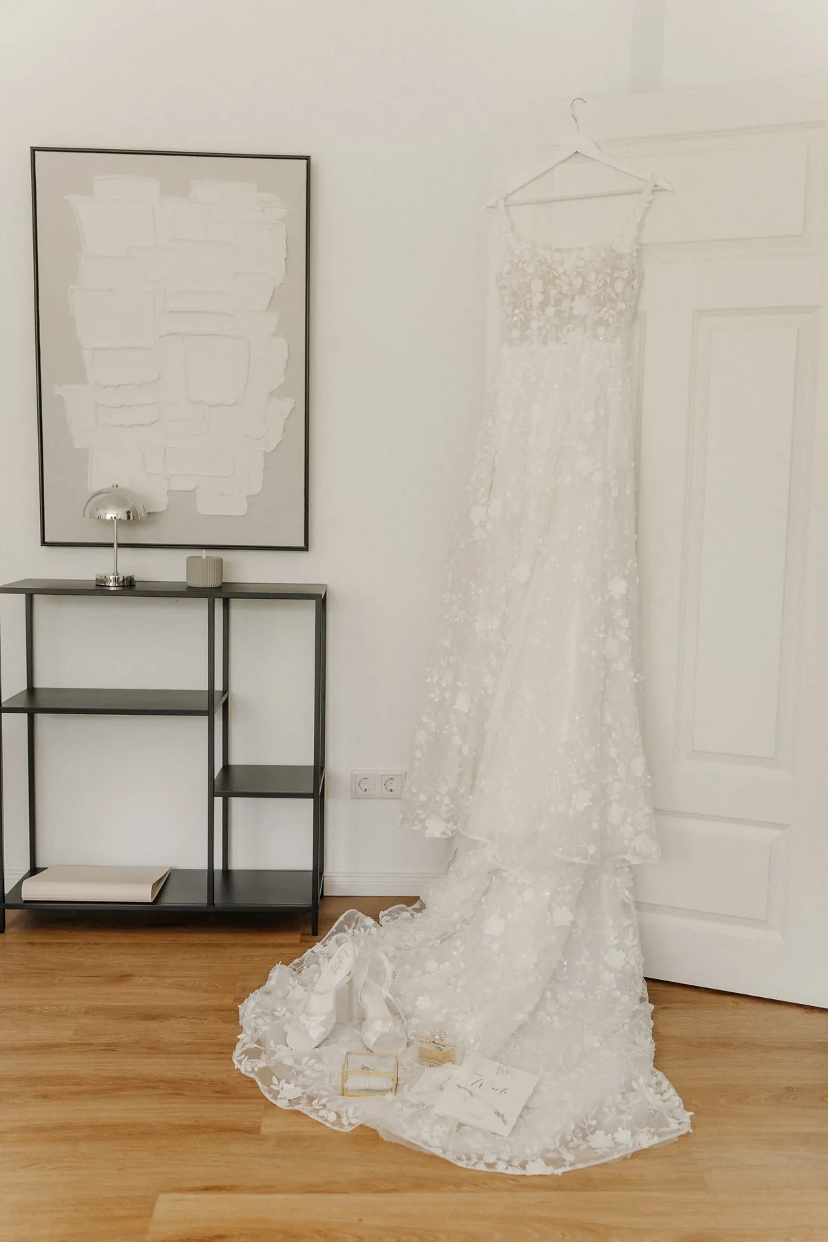 Das Brautkleid mit Blütenspitze hängt an einer weißen Kassettentür neben einem schwarzen Metallregal, die lange Schleppe breitet sich über den Holzboden, darauf Brautschuhe, Ringkästchen, Parfüm und ein Büchlein.
