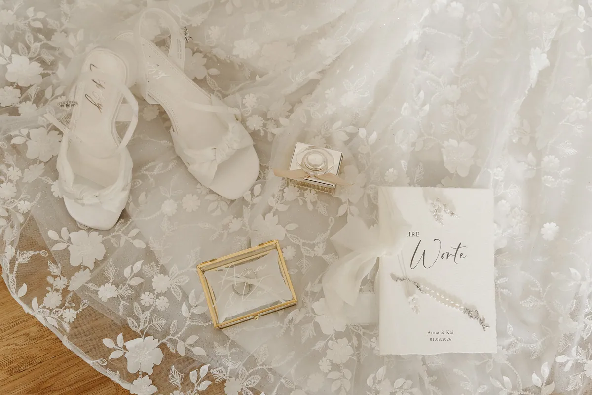 Von oben auf der Spitzenschleppe arrangiert: weiße Sandaletten mit Knotendetail, ein Parfümflakon, das goldgerahmte Ringkästchen mit „Anna & Kai“ und ein Büchlein „Ihre Worte“ mit Perlenschmuck.