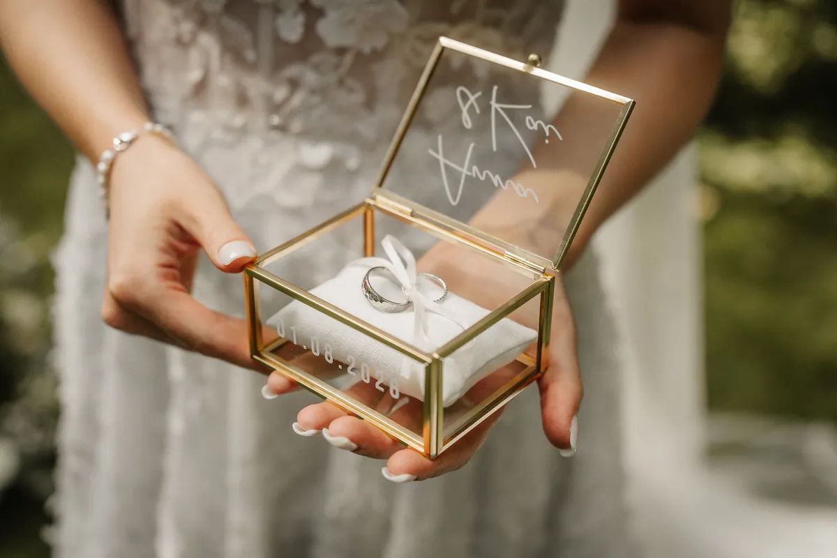 Annas Hände halten ein goldgerahmtes Glaskästchen mit zwei Ringen auf weißem Kissen, im Deckel steht in Schreibschrift „Anna & Kai“, auf der Front das Datum 01.08.2026.