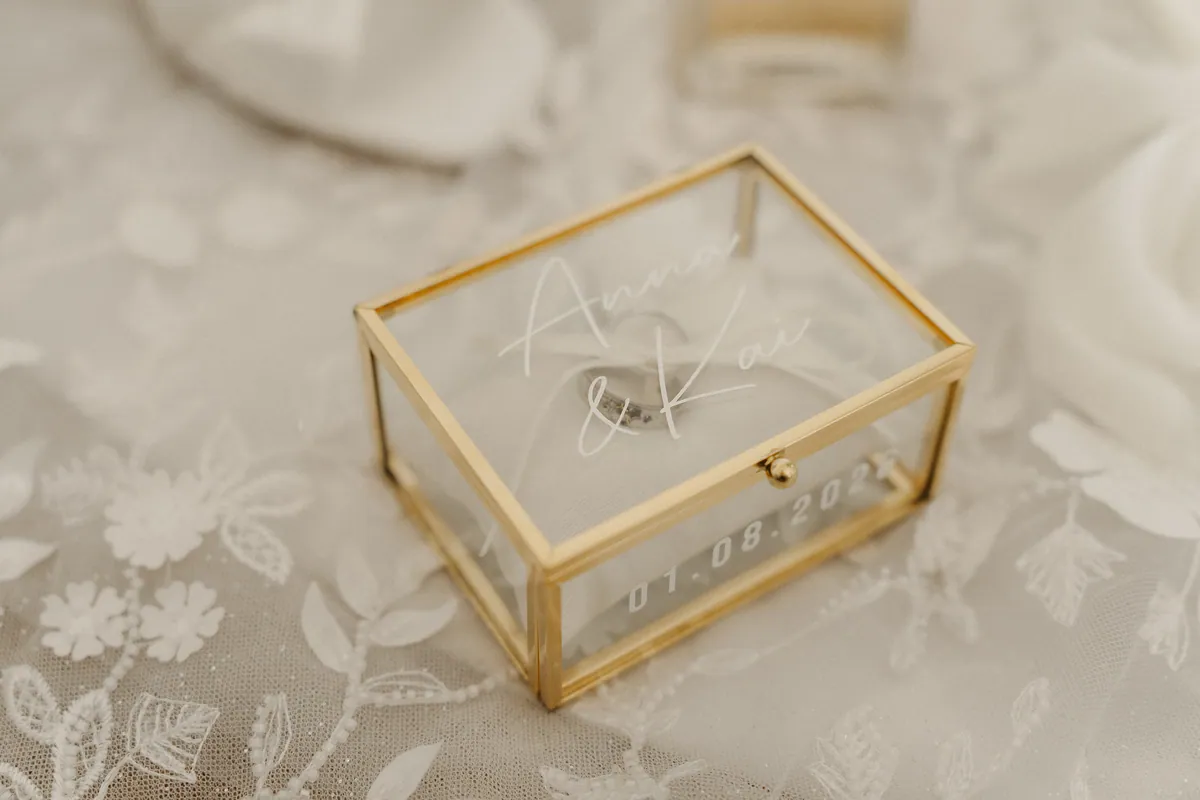 Ein Glaskästchen mit Goldrahmen steht auf der Brautkleidspitze, auf dem Deckel in weißer Schreibschrift „Anna & Kai“, an der Seite „01.08.2026“, darunter schimmert ein Ring.