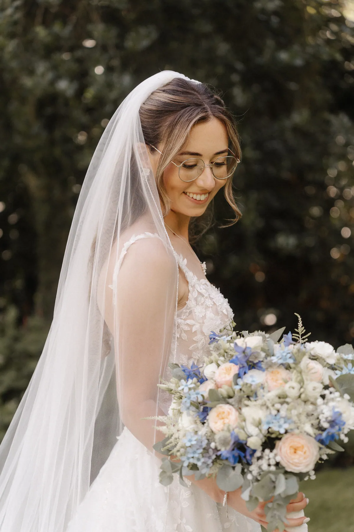 Anna blickt lächelnd auf ihren Strauß aus pfirsichfarbenen Rosen, tiefblauen und hellblauen Blüten und Eukalyptus, der lange Schleier fällt ihr über den Rücken.