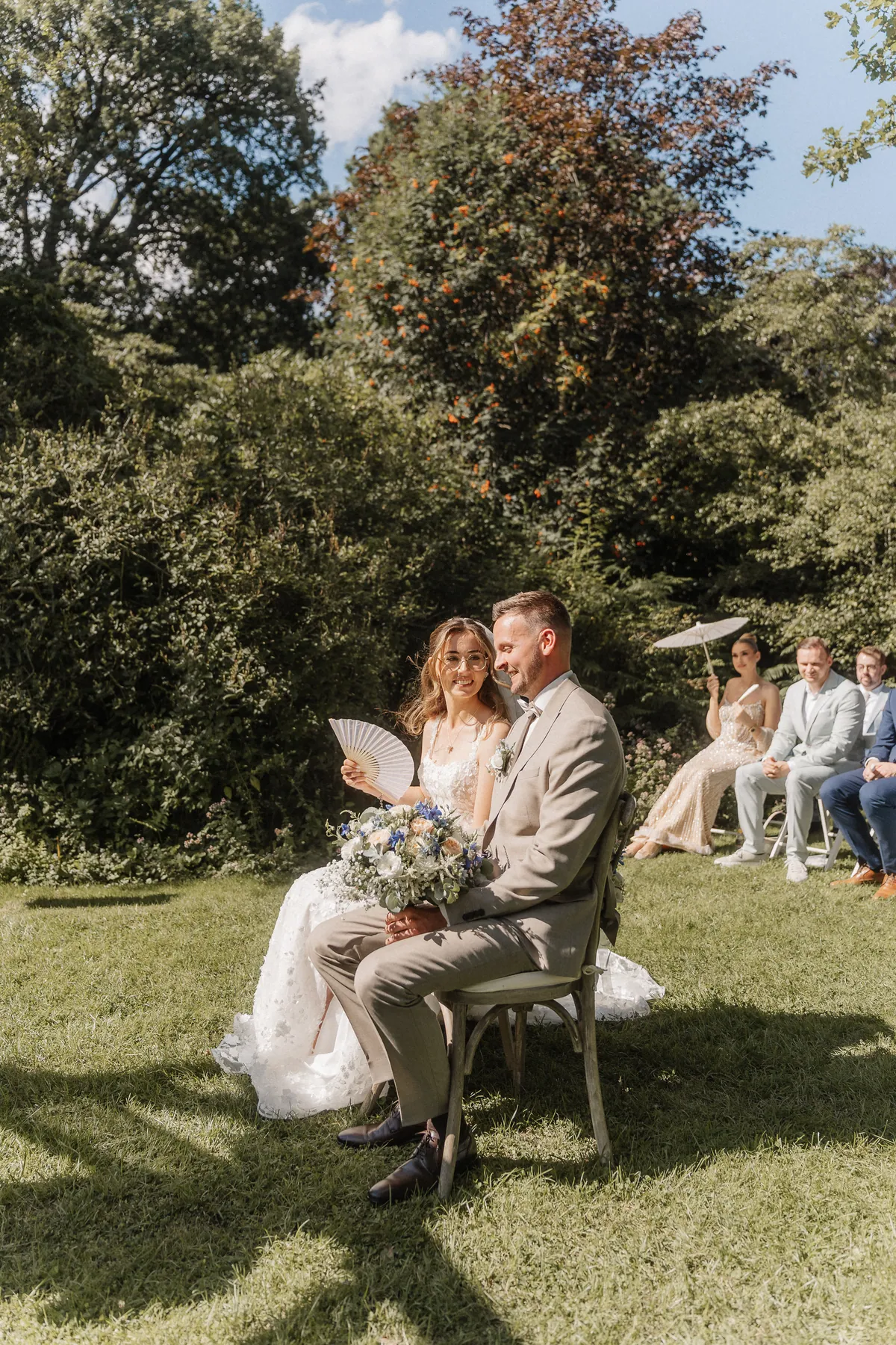 Anna und Kai sitzen auf Holzstühlen im Gras, sie fächelt sich mit einem weißen Papierfächer Luft zu und lächelt ihn an, den Strauß auf dem Schoß.