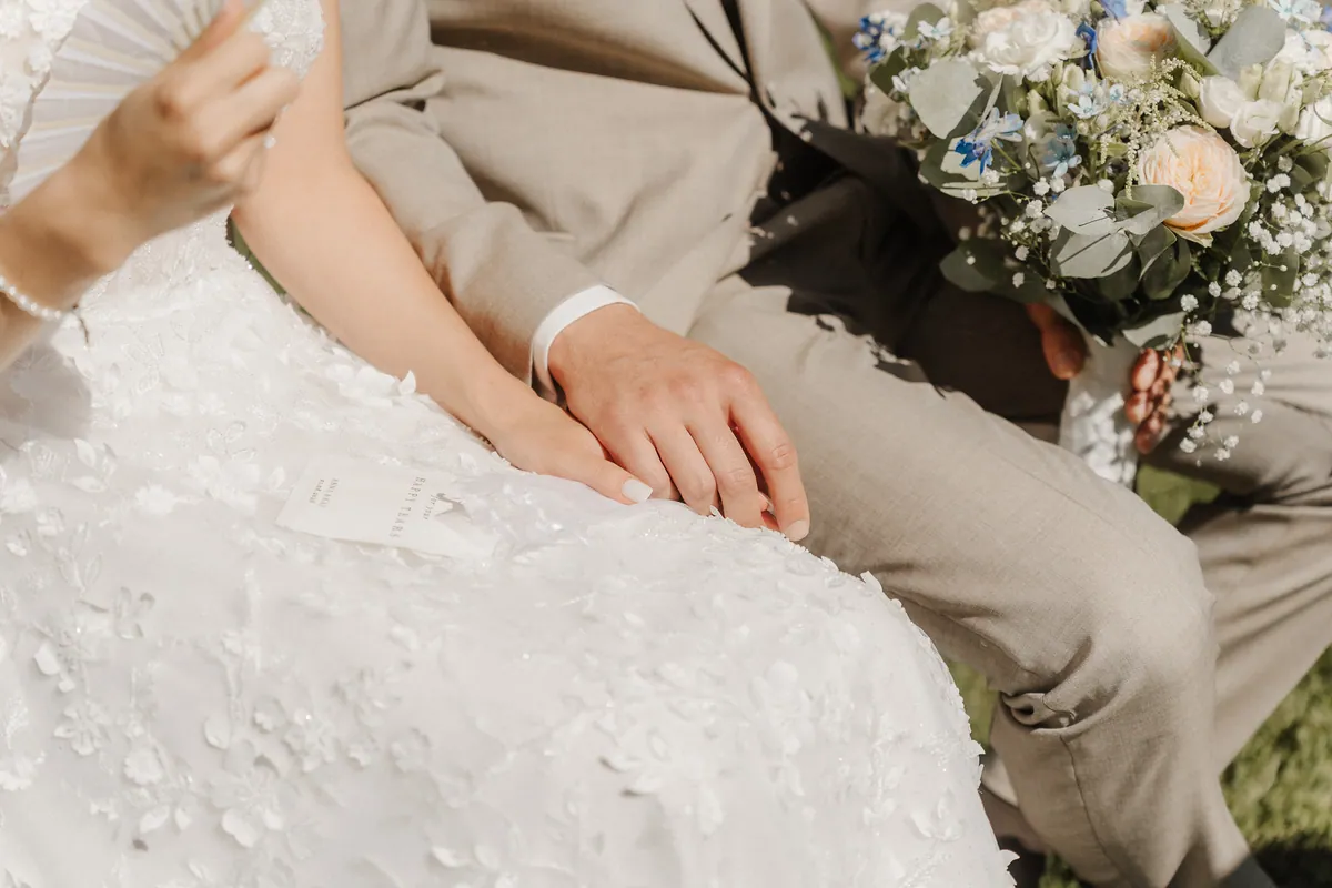 Nahaufnahme auf dem Schoß: Kais Hand liegt auf Annas, daneben ein Kärtchen mit der Aufschrift «for your happy tears», rechts der Strauß aus Pfirsichrosen, Schleierkraut und blauen Blüten.