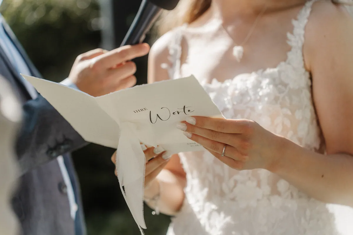 Nahaufnahme: Anna hält eine Karte aus Büttenpapier mit der Aufschrift «Ihre Worte» und weißem Stoffband, am Ringfinger ein schmaler Ring mit kleinem Stein, von links hält eine Hand das Mikrofon.