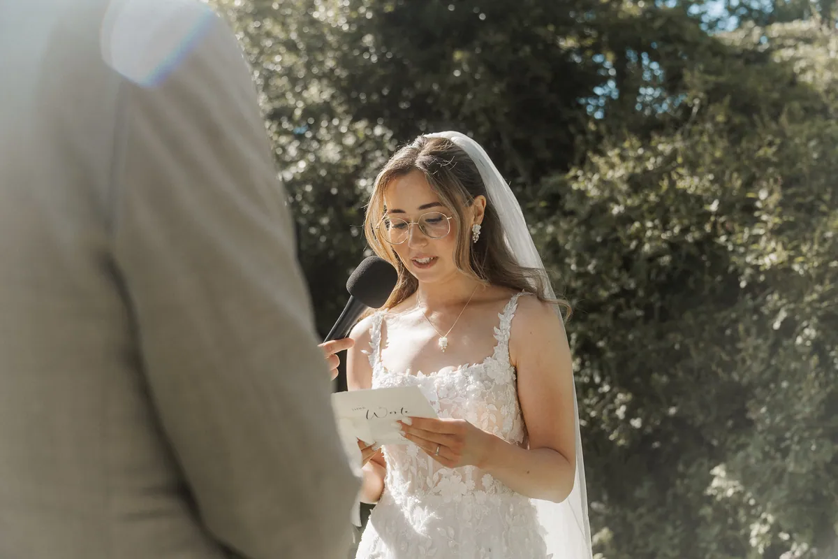 Anna liest mit Brille und Schleier von ihrer Karte ab und spricht in ein Mikrofon, das ihr von links hingehalten wird, im Vordergrund unscharf Kais Schulter im hellen Sakko.