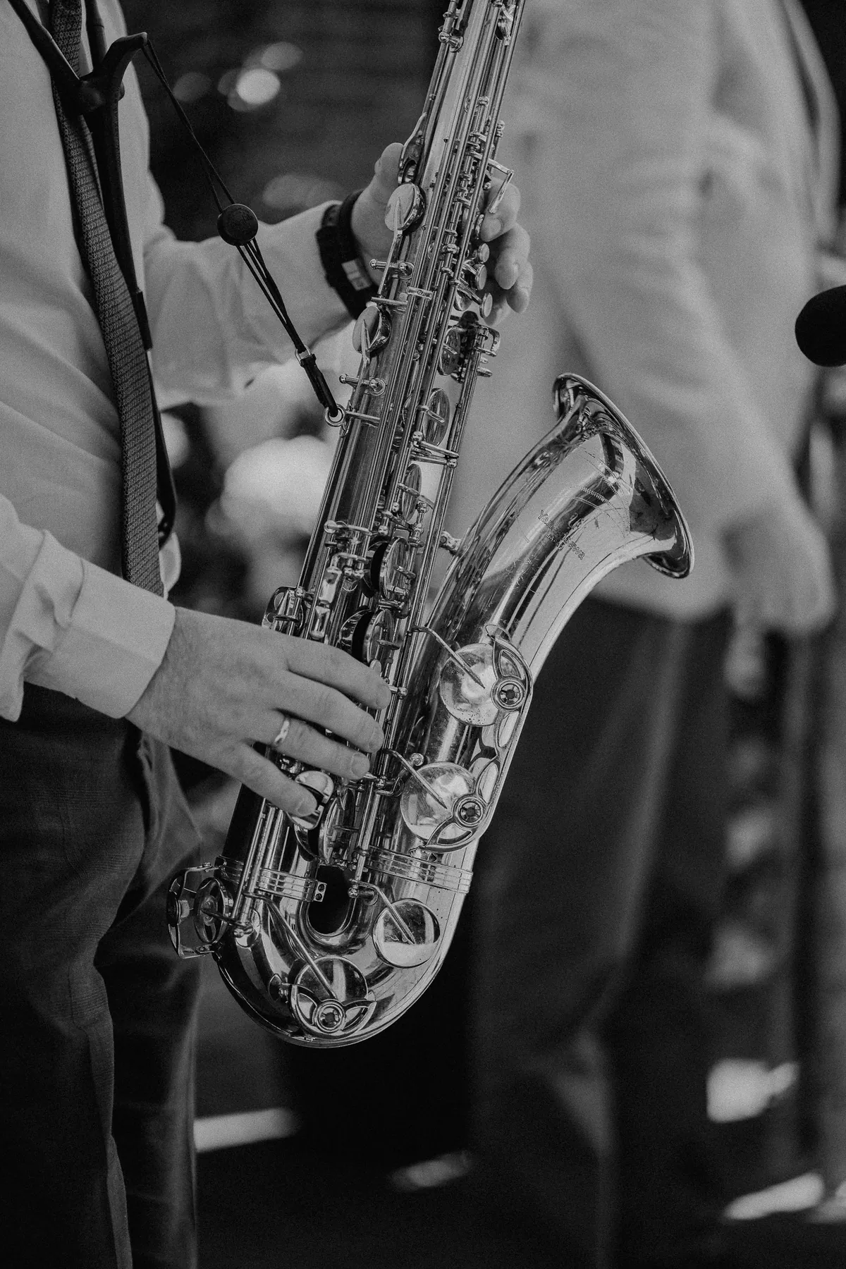 In Schwarzweiß: Nahaufnahme eines metallisch glänzenden Saxofons, das ein Musiker in hellem Hemd und gemusterter Krawatte mit beiden Händen hält, am Hals ein Tragegurt, rechts unscharf ein Mikrofon.