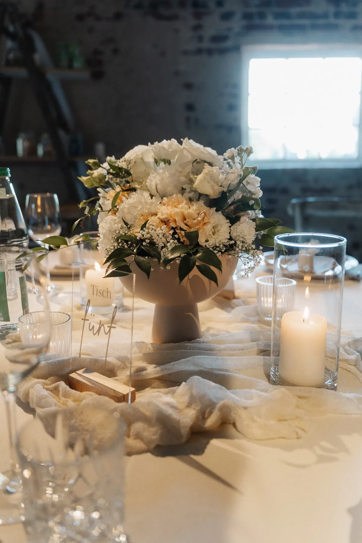 Auf dem gedeckten Tisch steht eine Fußschale mit weißen und apricotfarbenen Blüten, daneben brennende Stumpenkerzen im Glas und ein Acrylschild mit der Aufschrift «Tisch fünf».