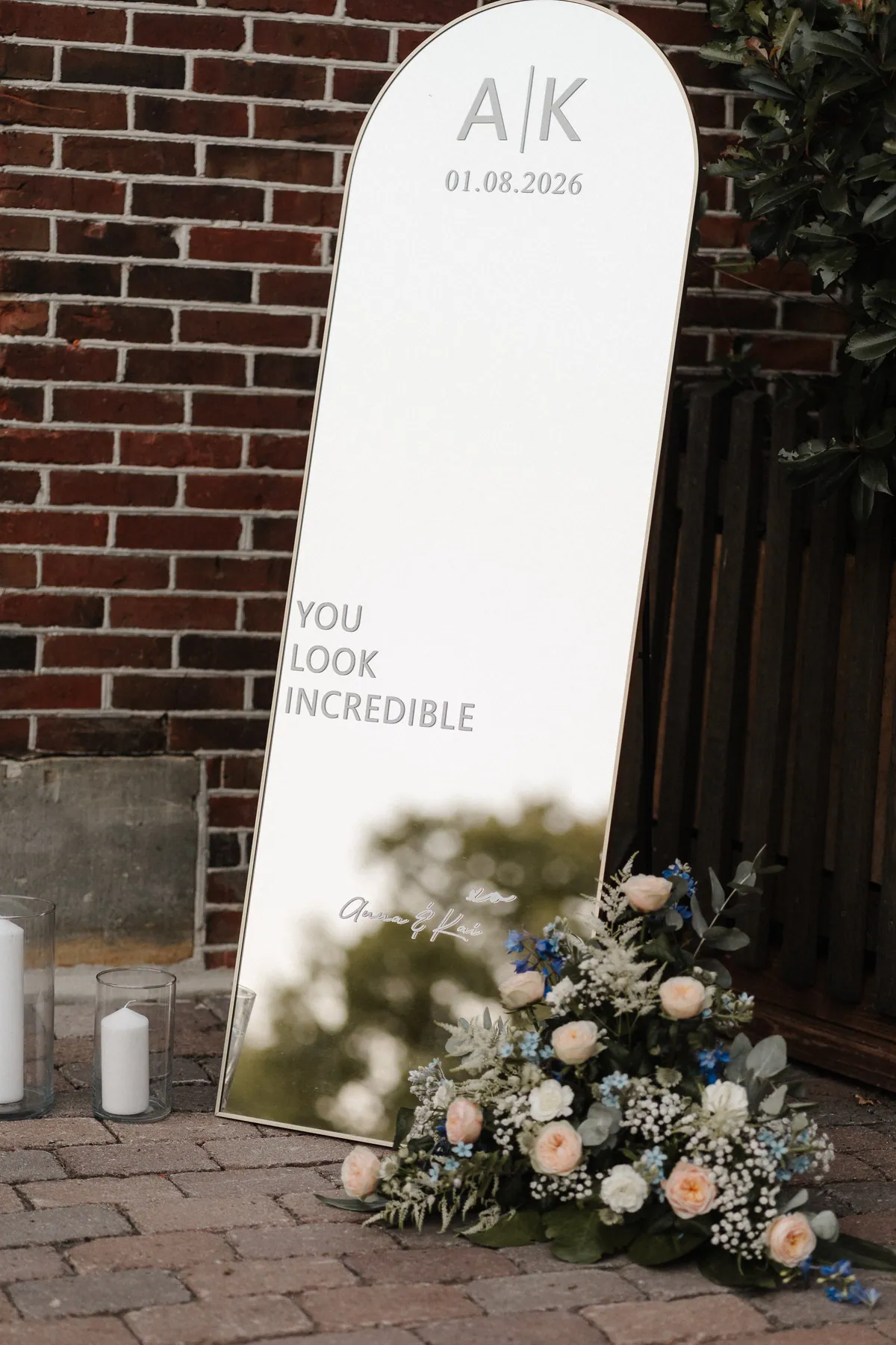 Ein bogenförmiger Spiegel lehnt an einer Backsteinwand, darauf «A|K 01.08.2026» und «You look incredible», unten der Schriftzug «Anna & Kai» und am Fuß ein üppiges Blumenarrangement.