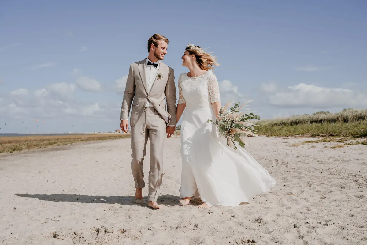 Barfuß und Hand in Hand laufen Braut und Bräutigam über den Sandstrand und lachen sich an, ihr Kleid weht, im Arm ein Strauß mit Pampasgras.