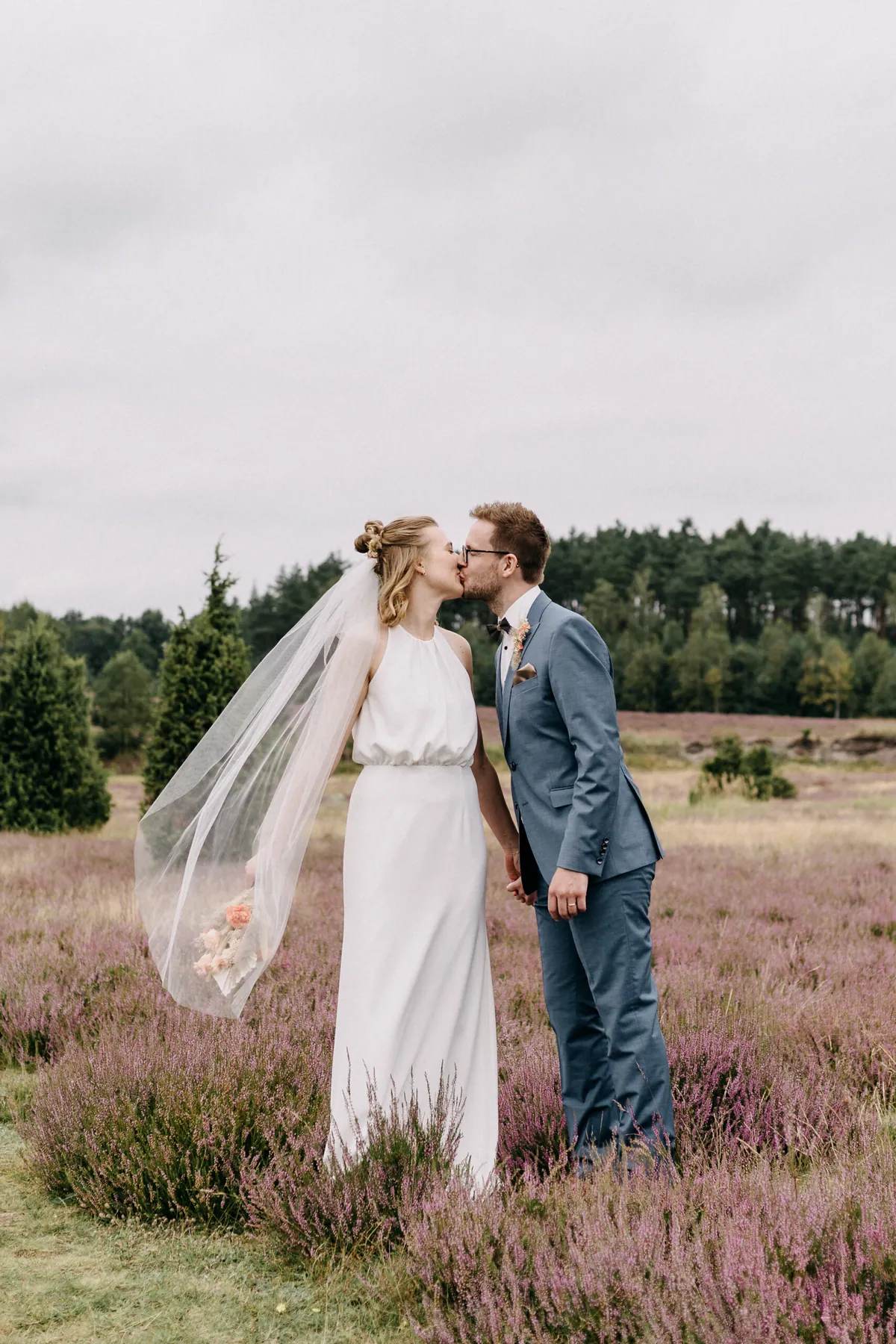 Das Brautpaar küsst sich inmitten lila blühender Heide, der lange Schleier weht im Wind, dahinter Kiefern unter grauem Himmel.