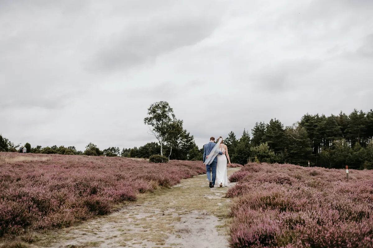 Eng umschlungen geht das Brautpaar einen sandigen Weg durch die Heide am Pestruper Gräberfeld entlang, darüber graue Wolken.