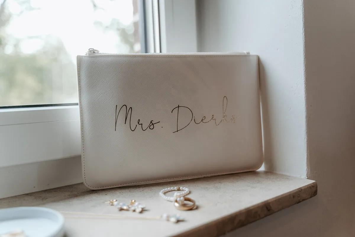 Auf der Fensterbank lehnt eine cremeweiße Clutch mit goldener «Mrs.»-Aufschrift, davor liegen Ohrringe, ein Perlenarmband, ein Ring und eine Kette.