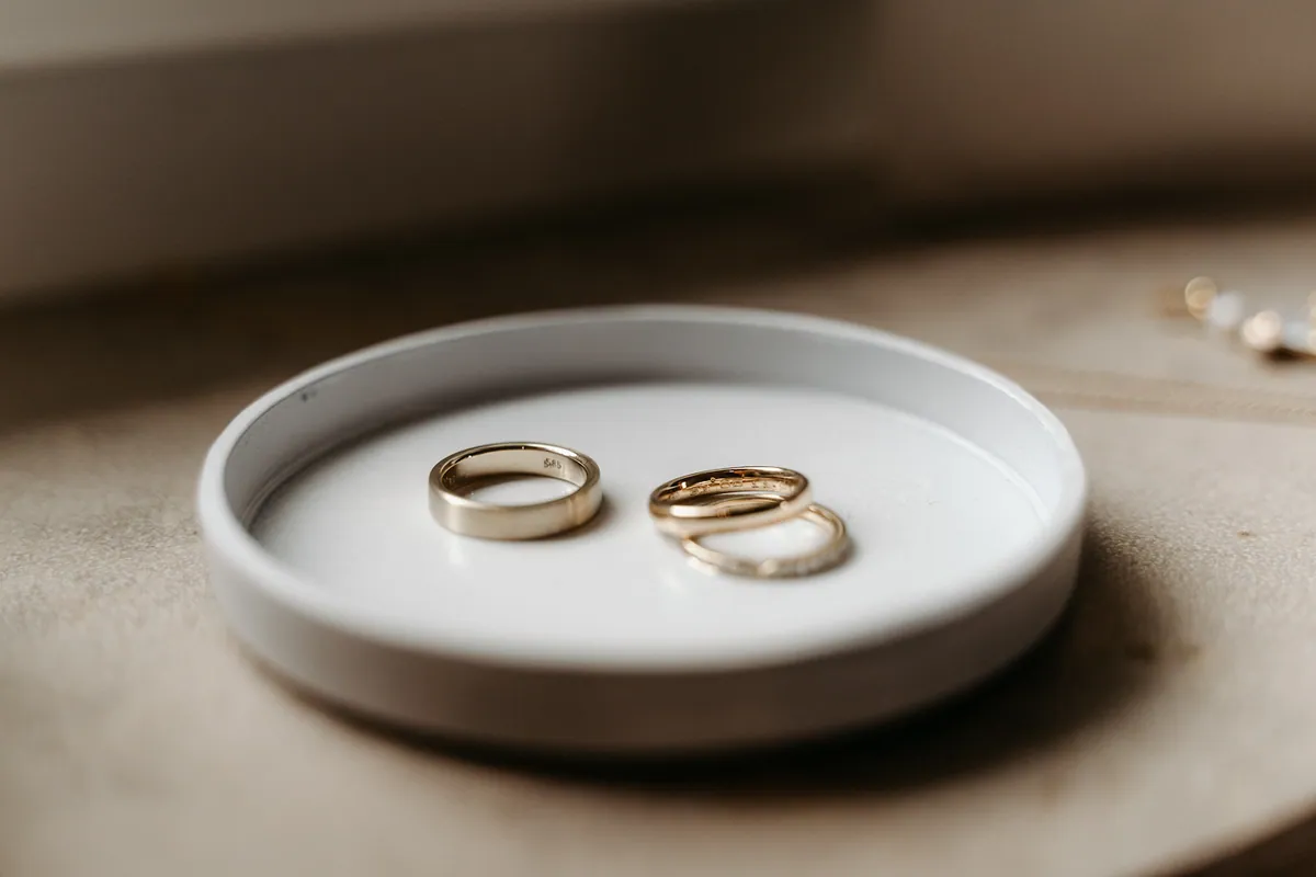 Die Eheringe liegen in einer kleinen weißen Keramikschale: ein schlichter goldener Ring und daneben zwei schmale, übereinanderliegende Ringe.