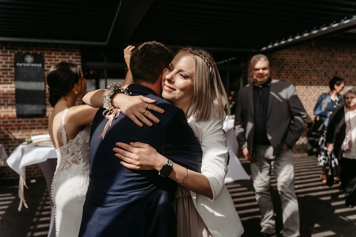 Eine blonde Freundin im weißen Blazer umarmt den Bräutigam herzlich, im Hintergrund stehen die Braut und lächelnde Gäste im Sonnenlicht.