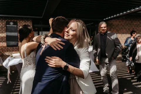 Eine blonde Freundin im weißen Blazer umarmt den Bräutigam herzlich, im Hintergrund stehen die Braut und lächelnde Gäste im Sonnenlicht.