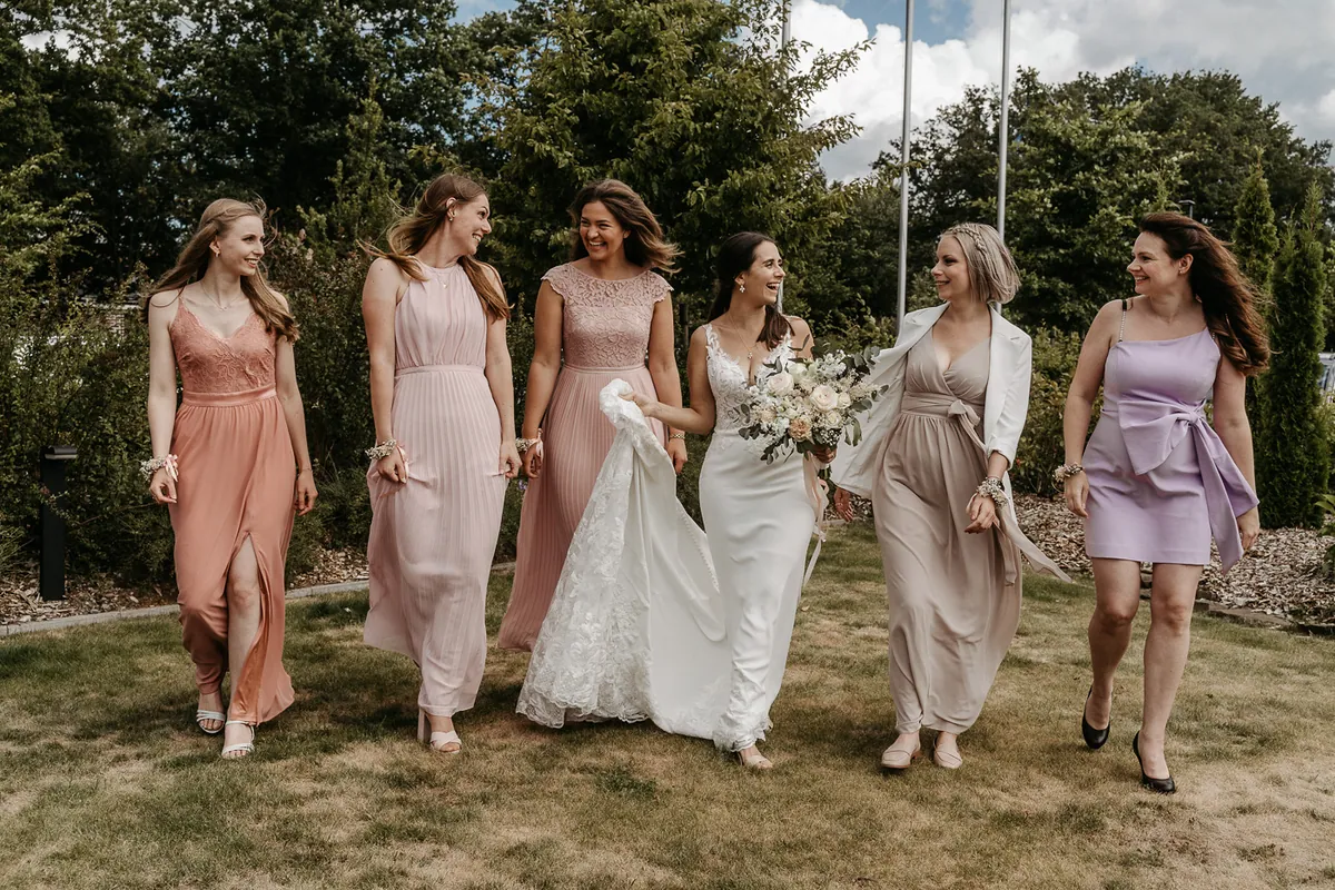 Lachend läuft die Braut mit fünf Freundinnen in Kleidern in Rosé, Nude und Flieder über eine sommerliche Wiese.