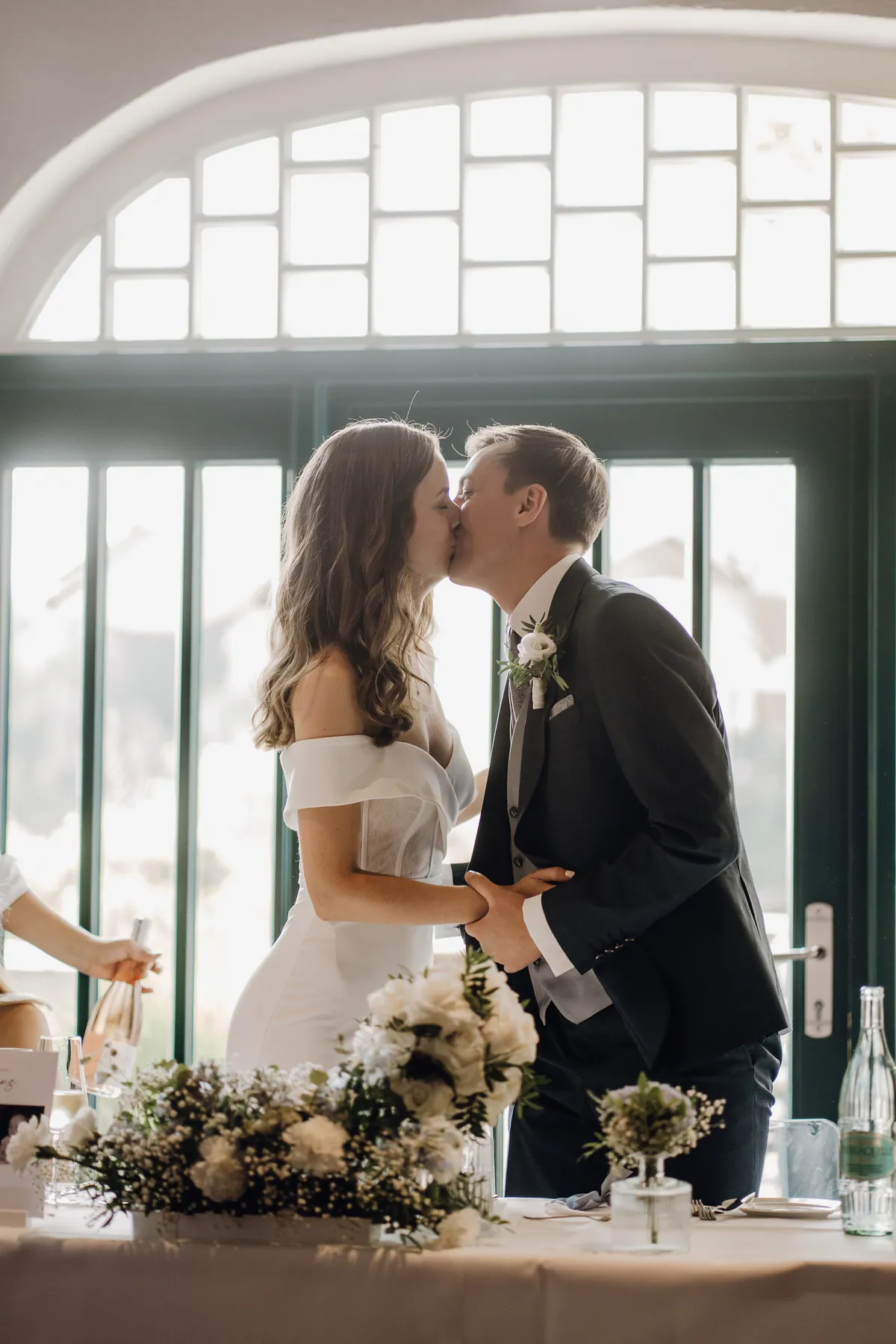 Im Gegenlicht küssen sich Luisa und Jonas am Brauttisch vor grün gerahmten Glastüren unter einem Rundbogenfenster, sie halten sich an den Händen, vorne weiße Blumen.