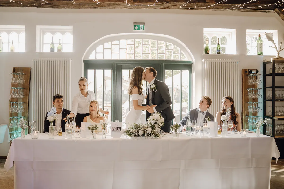Am langen weißen Brauttisch küssen sich Luisa und Jonas vor der Rundbogentür, links schenkt eine Servicekraft einem Gast Wasser ein, rechts blicken zwei Gäste lächelnd zu ihnen hoch.