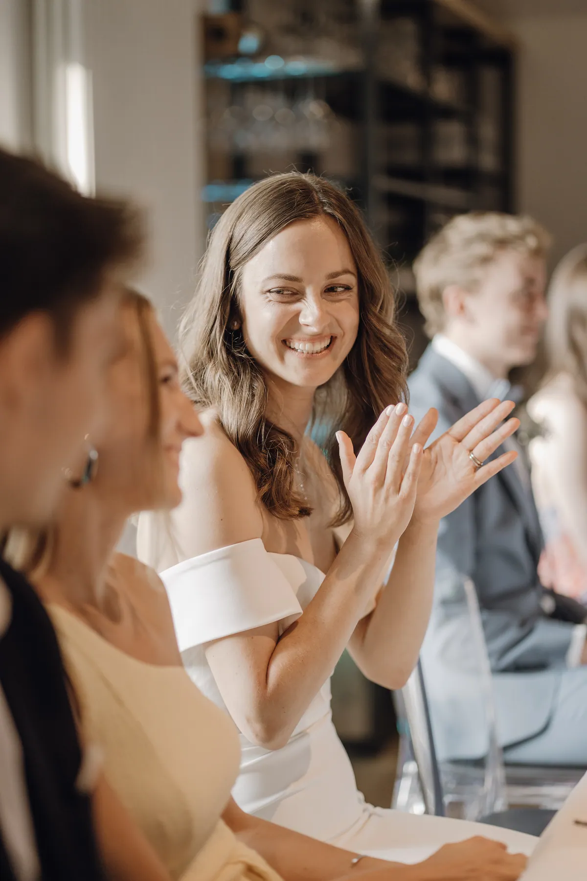 Luisa sitzt an der Tafel, klatscht in die Hände und lacht zur Seite, am Ringfinger ein schmaler Ring, vorne unscharf zwei Gäste, hinten ein Regal mit Gläsern.