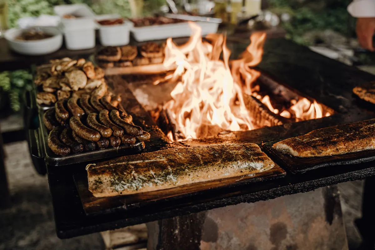 Auf einer Feuerplatte lodern Flammen zwischen Lachsfilets auf Holzbrettern, Bratwürsten, hellem Grillfleisch und Fleischpatties, dahinter Schalen mit Beilagen.