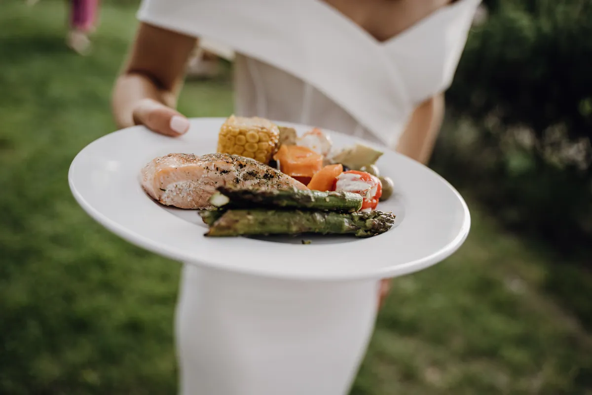 Nah am Teller in Luisas Händen: gegrillter Lachs, grüner Spargel, ein Stück Maiskolben, Möhren, gefüllte Paprika und Oliven, dahinter das weiße schulterfreie Kleid.