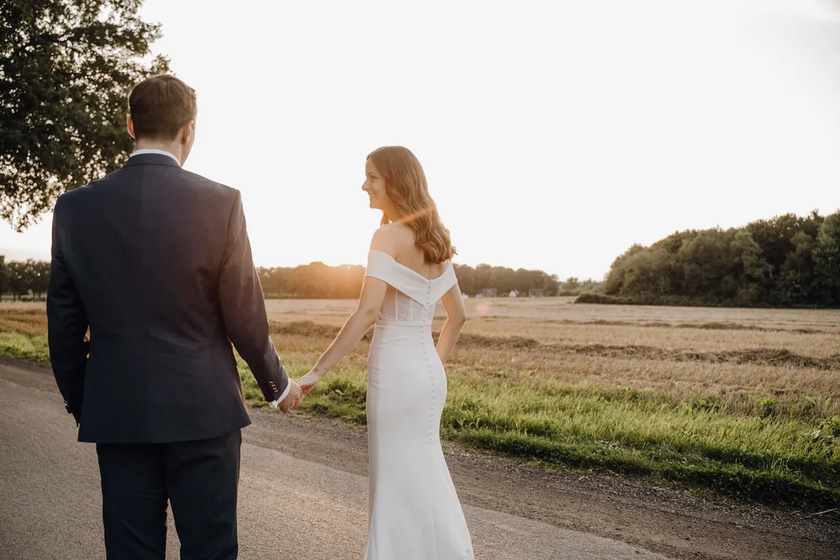 Von hinten gesehen halten sich Luisa und Jonas auf einer schmalen Straße am abgeernteten Feld an der Hand, sie blickt lächelnd zur Seite, über den Bäumen steht tief die Sonne.