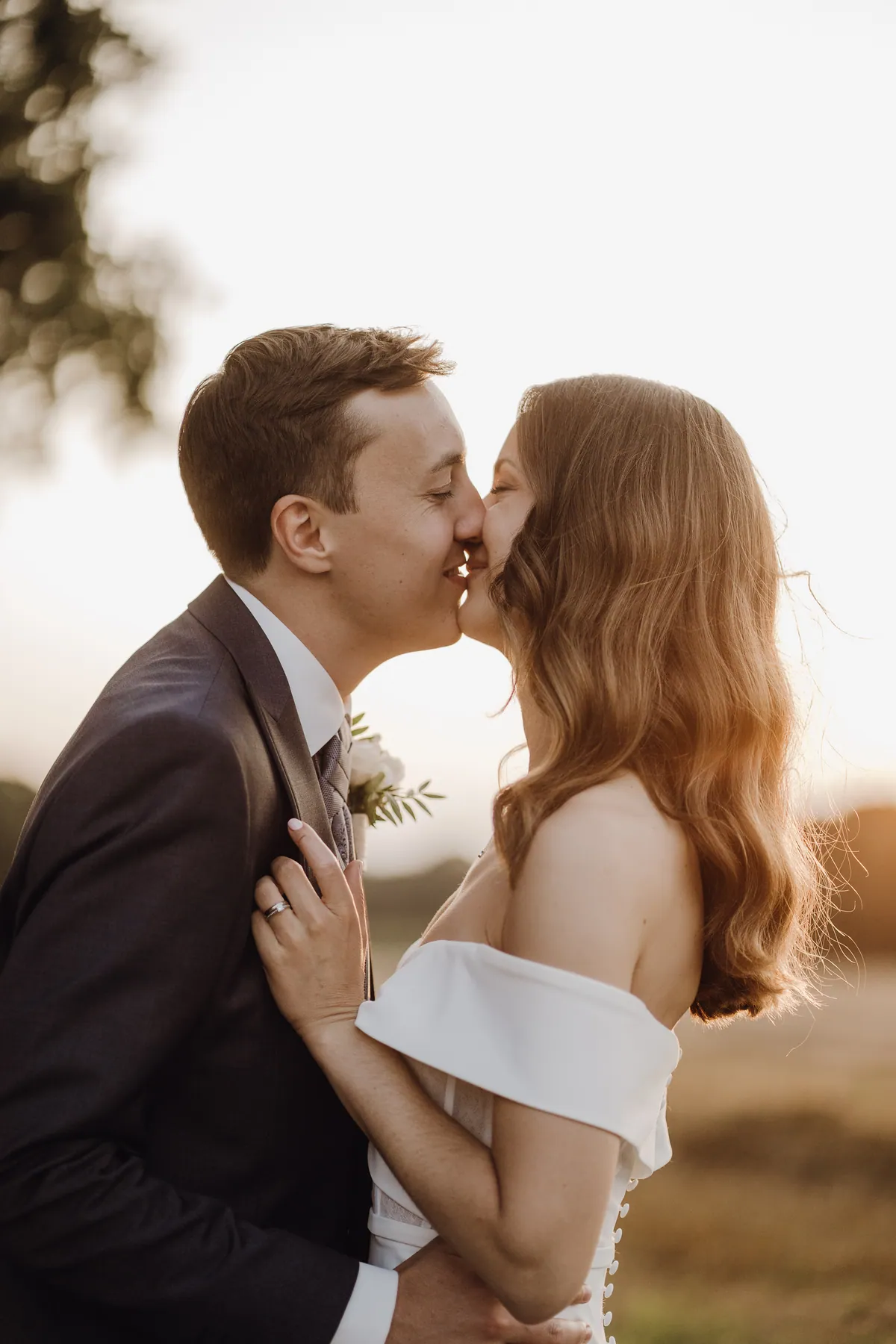 Im warmen Gegenlicht küssen sich Luisa und Jonas lächelnd mit geschlossenen Augen, ihre Hand mit dem Ring liegt auf seinem Revers unterhalb der weißen Ansteckblume, ihr langes gewelltes Haar leuchtet.
