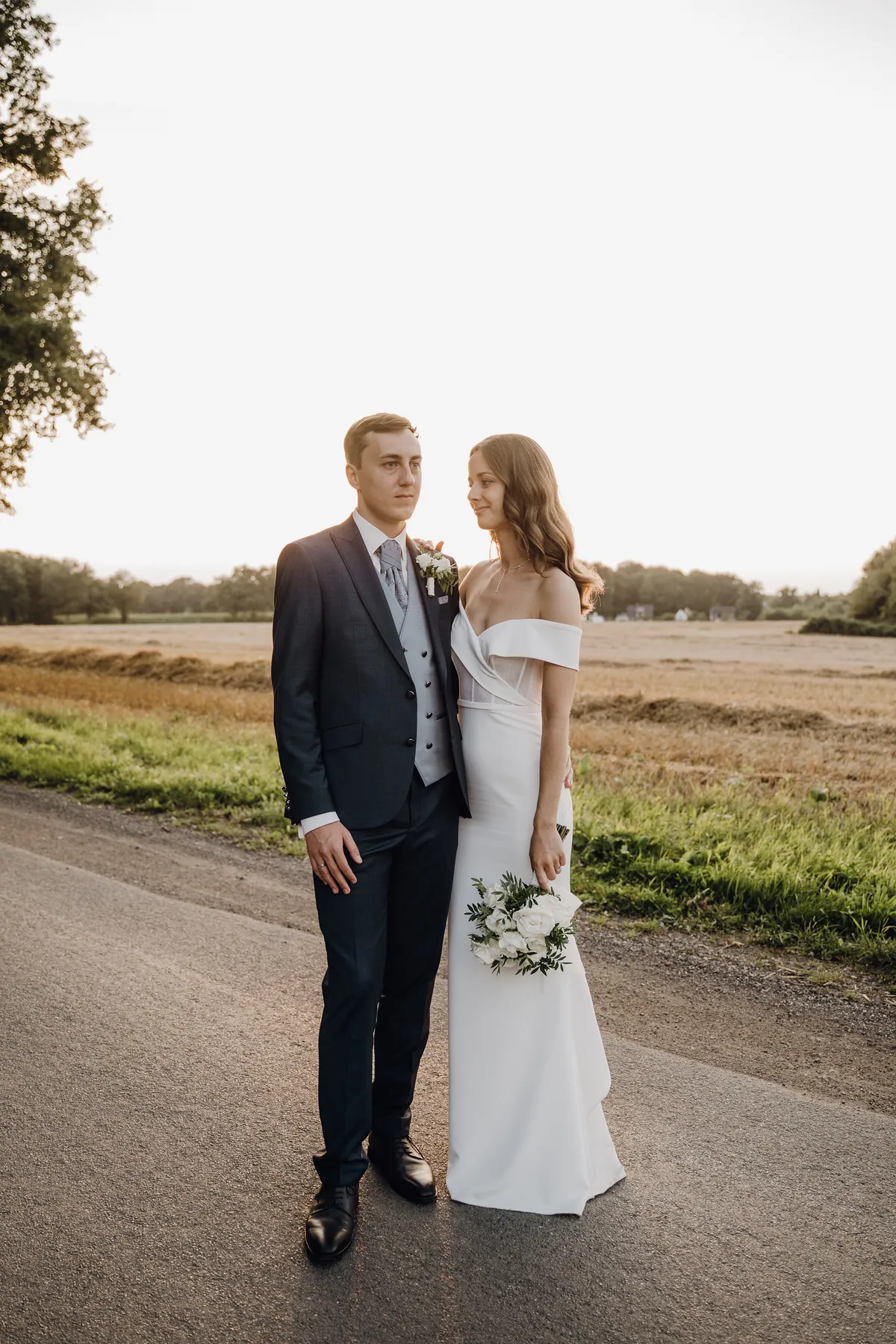 Luisa und Jonas stehen eng nebeneinander auf der Landstraße, er blickt in die Ferne, sie hat eine Hand auf seiner Schulter und schaut lächelnd zu ihm, in der anderen der Strauß aus weißen Rosen.