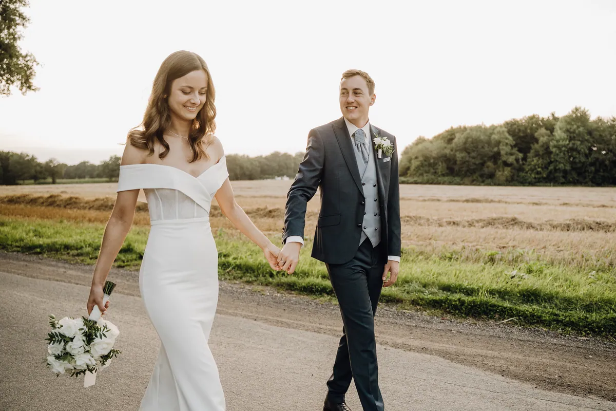 Hand in Hand spazieren Luisa und Jonas auf der Landstraße am Feld entlang, sie blickt lächelnd nach unten, er lächelt zu ihr hinüber, in ihrer anderen Hand baumelt der Strauß aus weißen Rosen.