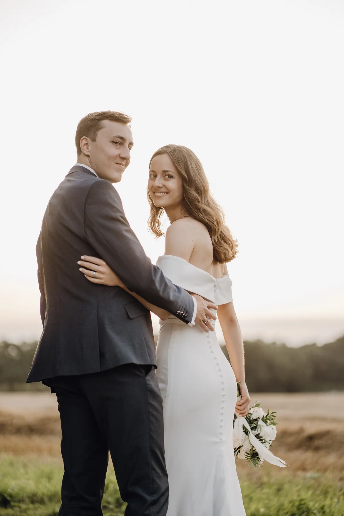 Von hinten gesehen halten sich Luisa und Jonas im Arm und blicken lächelnd über die Schulter in die Kamera, seine beringte Hand liegt an ihrer Taille, in ihrer Hand der Strauß mit herabhängendem Band.