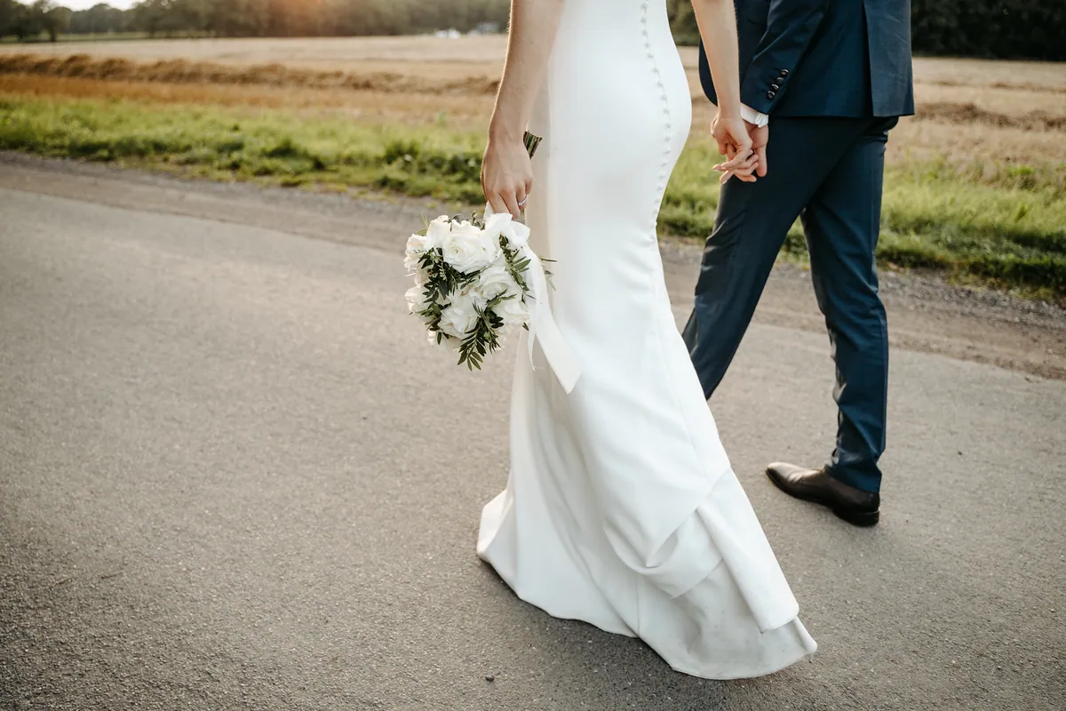 Von hinten, ab der Taille abwärts: Luisa trägt den Strauß aus weißen Rosen und Grün in der beringten Hand, mit der anderen hält sie Jonas’ Hand, die Schleppe gleitet über den Asphalt.