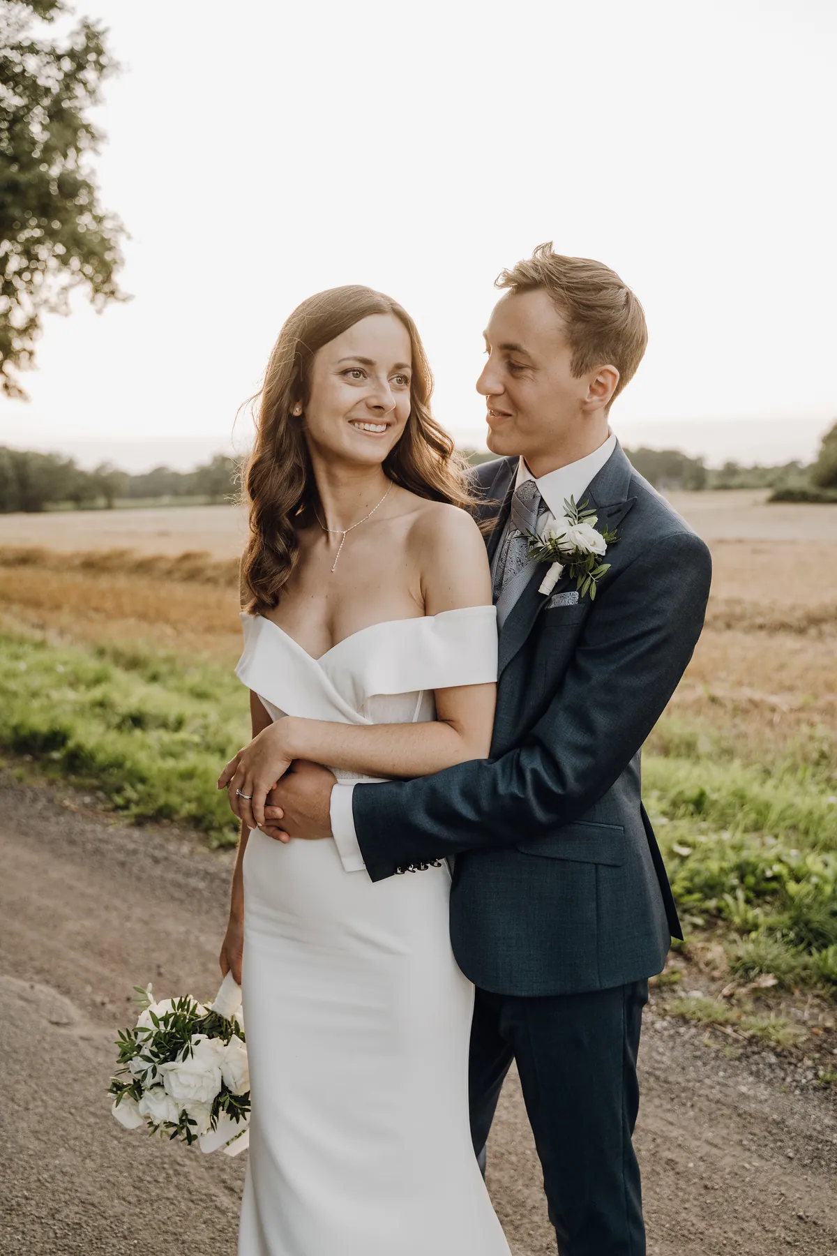 Jonas umarmt Luisa von hinten und schaut sie lächelnd an, sie legt ihre Hand mit dem Ring auf seinen Arm und lächelt breit zur Seite, in der anderen hält sie den weißen Strauß.