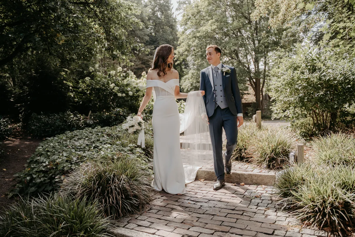 Luisa und Jonas gehen im Garten eine flache Stufe des Klinkerwegs hinab und sehen sich lächelnd an, sie trägt den Rosenstrauß in der einen und ein Stück Schleier in der anderen Hand.