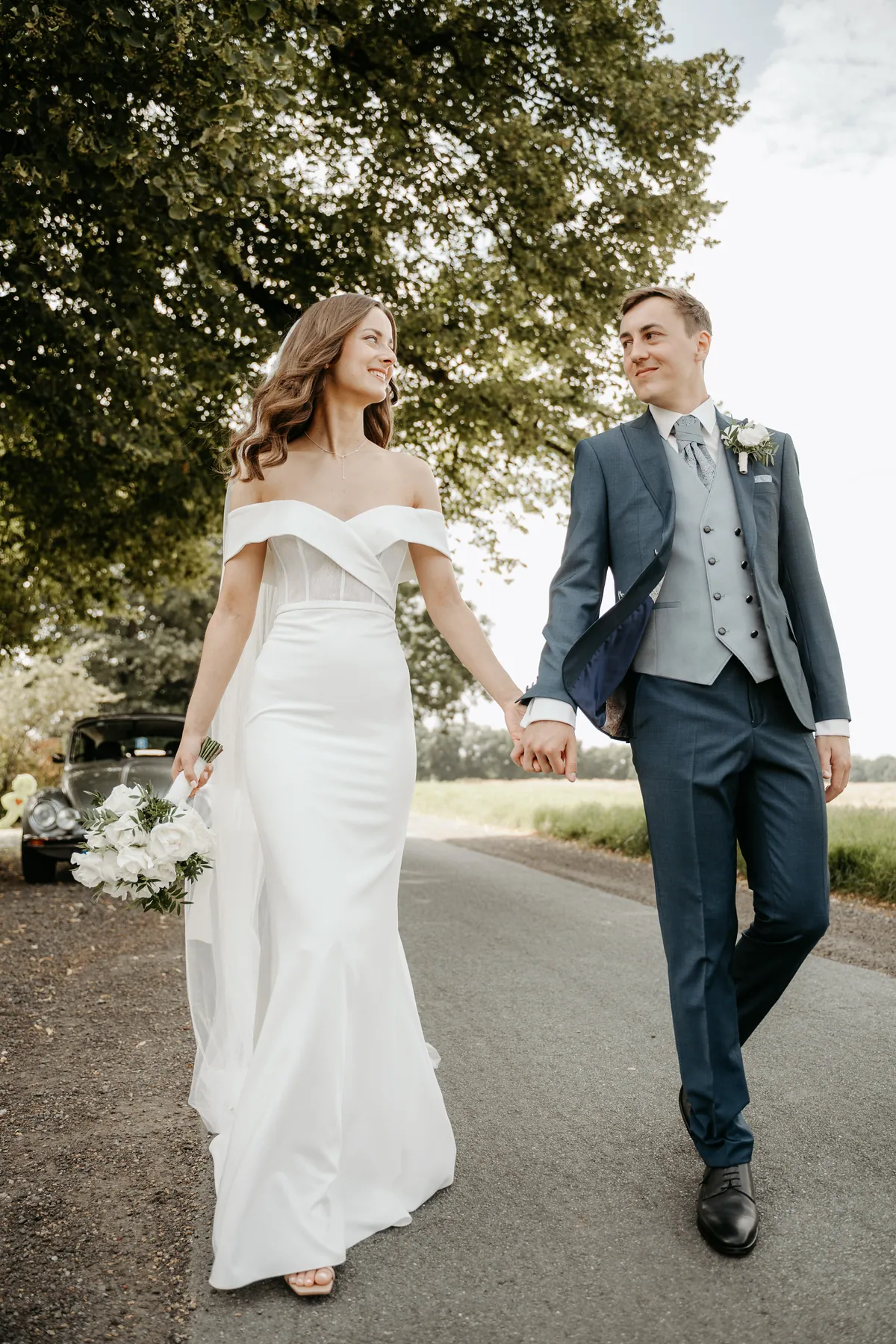 Luisa und Jonas gehen Hand in Hand die Landstraße entlang und schauen sich lächelnd an, sie trägt einen Strauß weißer Rosen, er eine hellgraublaue Weste unter dem dunkelblauen Sakko.