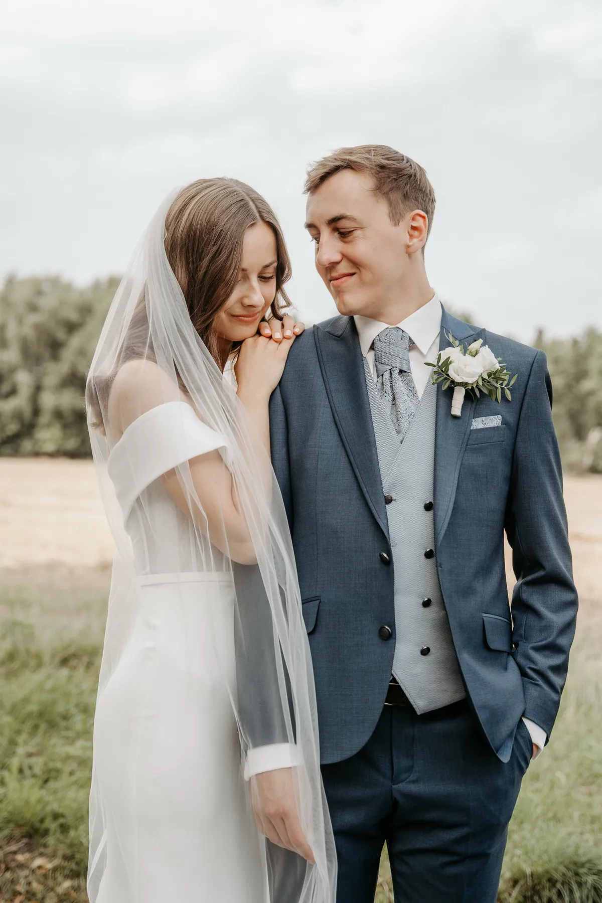 Luisa legt Kinn und Hände auf Jonas' Schulter und senkt lächelnd den Blick, der Schleier fällt über ihren Arm, Jonas schaut mit der Hand in der Hosentasche zu ihr hinunter.
