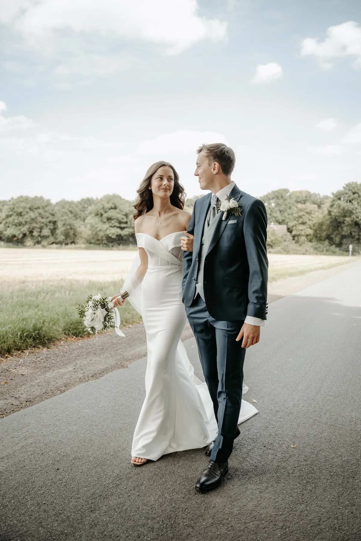 Eingehakt gehen Luisa und Jonas die Landstraße entlang und sehen sich an, neben ihnen ein abgeerntetes Feld und eine Baumreihe, darüber blauer Himmel mit weißen Wolken.