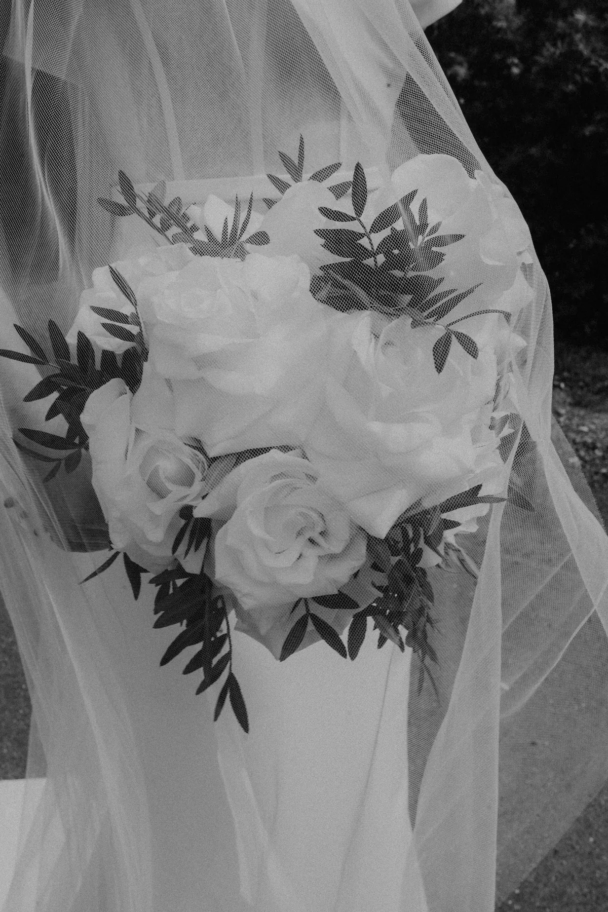 In Schwarzweiß: Der Strauß aus weißen Rosen und gefiederten Blättern, gesehen durch den Schleier, dessen feiner Tüll ein zartes Netzmuster über die Blüten legt.