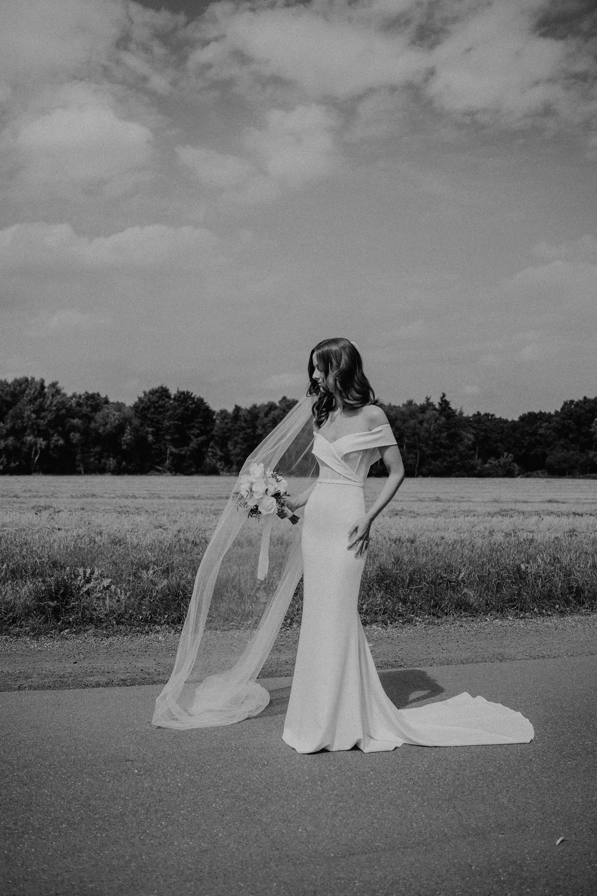 Schwarzweißaufnahme: Luisa steht allein auf der Straße, eine Hand an der Hüfte, und blickt über die Schulter nach unten, ihr Schleier bauscht sich zur Seite, dahinter Feld, Baumreihe und Wolkenhimmel.