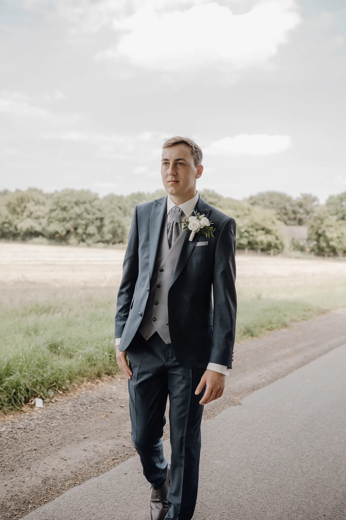 Jonas geht allein die Landstraße entlang und schaut zur Seite, im dunkelblauen Sakko mit hellgraublauer Weste, gemustertem Plastron und Ansteckblume, hinter ihm ein Feld und eine Baumreihe.