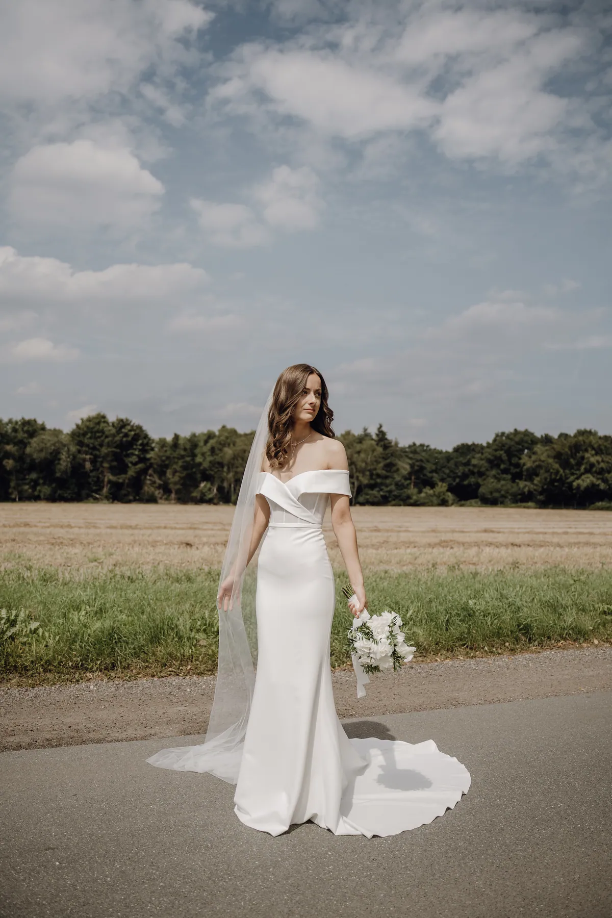 Luisa steht allein auf dem Asphalt vor einem abgeernteten Feld und blickt zur Seite, Schleier und Schleppe liegen ausgebreitet, in der Hand der Rosenstrauß, darüber ein weiter Wolkenhimmel.