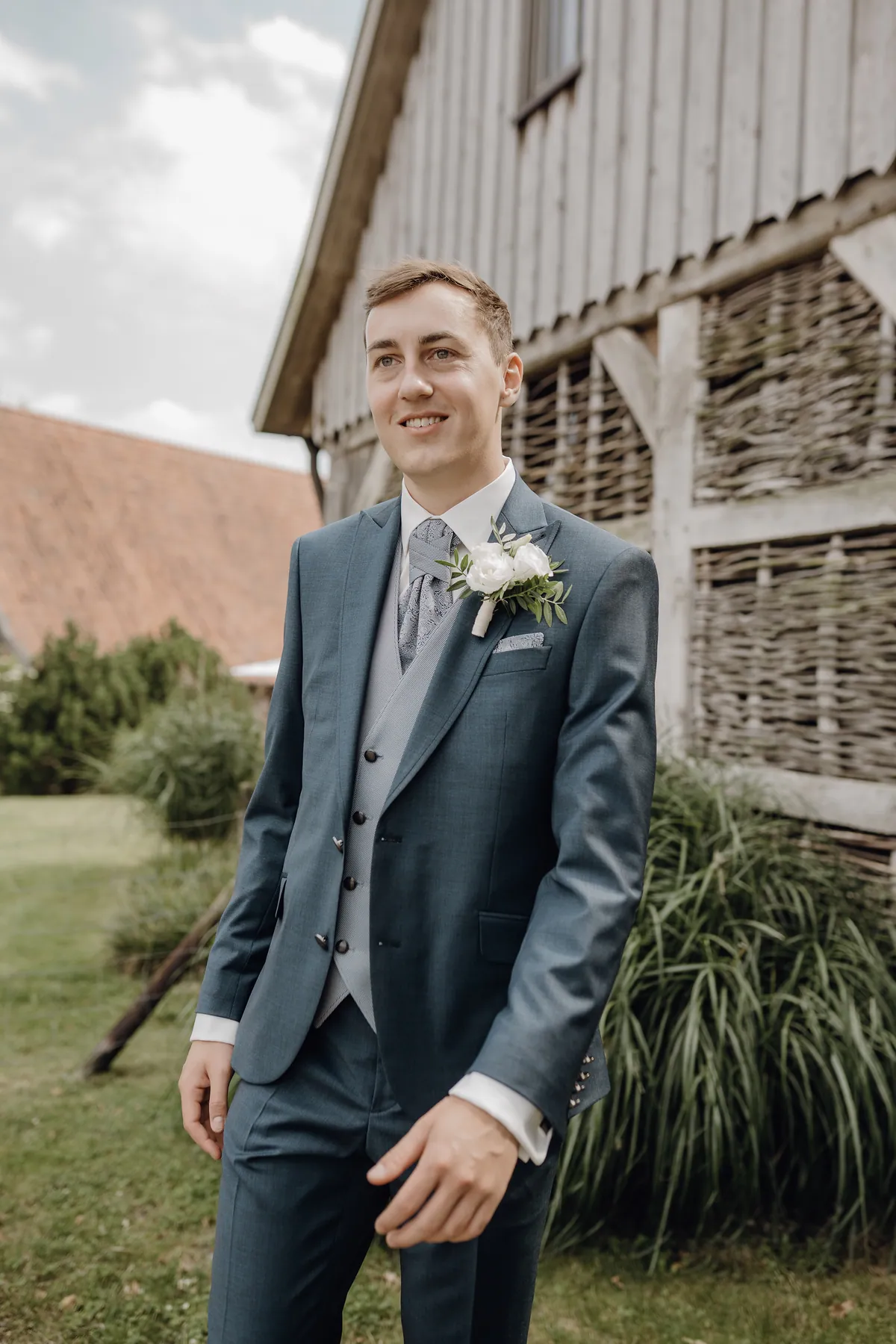 Im dunkelblauen Anzug mit weißer Ansteckblume lächelt Jonas zur Seite, hinter ihm ein Gebäude aus verwittertem Holz mit Flechtwerkwänden, hohe Gräser und ein rotes Ziegeldach.