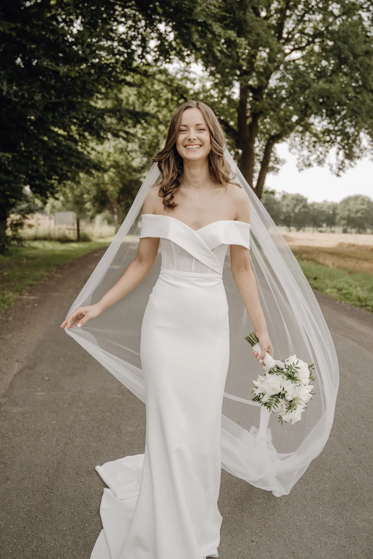 Auf einem Asphaltweg unter alten Bäumen lacht Luisa im schulterfreien Kleid in die Kamera, hält mit einer Hand den langen Schleier seitlich hoch, in der anderen weiße Rosen, rechts ein goldgelbes Feld.