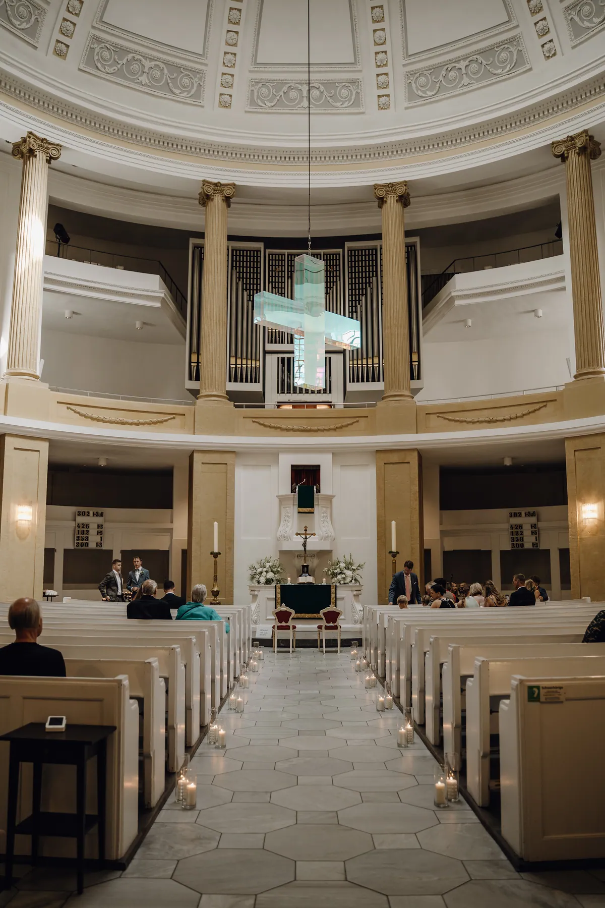 Blick durch den kerzengesäumten Mittelgang einer runden Kuppelkirche mit ionischen Säulen: am Ende zwei rote Stühle vor dem Altar, darüber hängt ein schimmerndes Glaskreuz vor der Orgel auf der Empore.